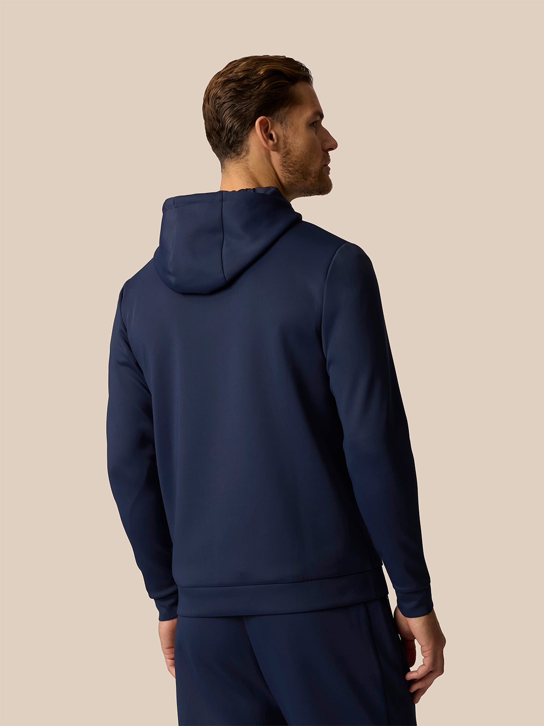 A man shown from the back wearing a navy blue hooded sweatshirt and matching pants against a solid beige background.