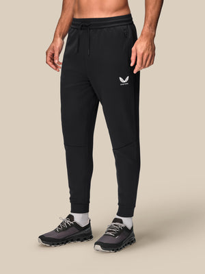 A person modeling black jogger-style sweatpants with a small white logo on the upper left thigh, shown from the waist down against a plain beige studio background.