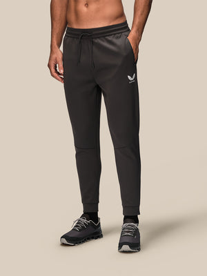 A man wearing dark gray athletic jogger pants with a drawstring waist and a small white logo on the left thigh, paired with dark running shoes, against a solid beige background.