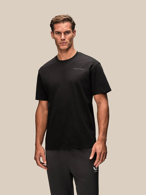 A man modeling a black short-sleeve crewneck t-shirt with a small white 'CASTORE' logo on the left chest, paired with dark gray athletic pants, against a solid beige background.