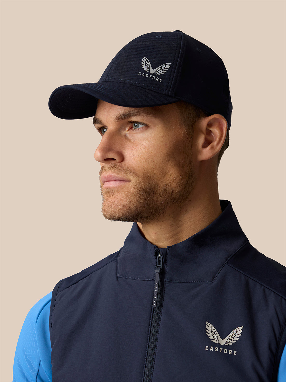 A man in profile wearing a navy blue Castore baseball cap and a matching navy blue zip-up vest over a bright blue shirt, against a solid beige background.