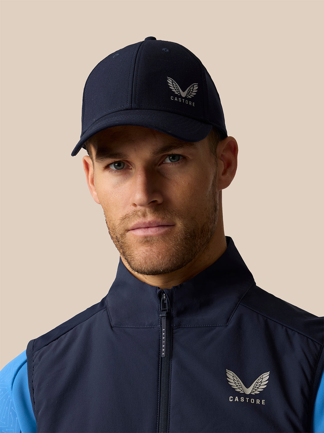 A man wearing a navy blue Castore baseball cap and a matching navy blue zip-up vest over a light blue shirt, set against a solid beige background.