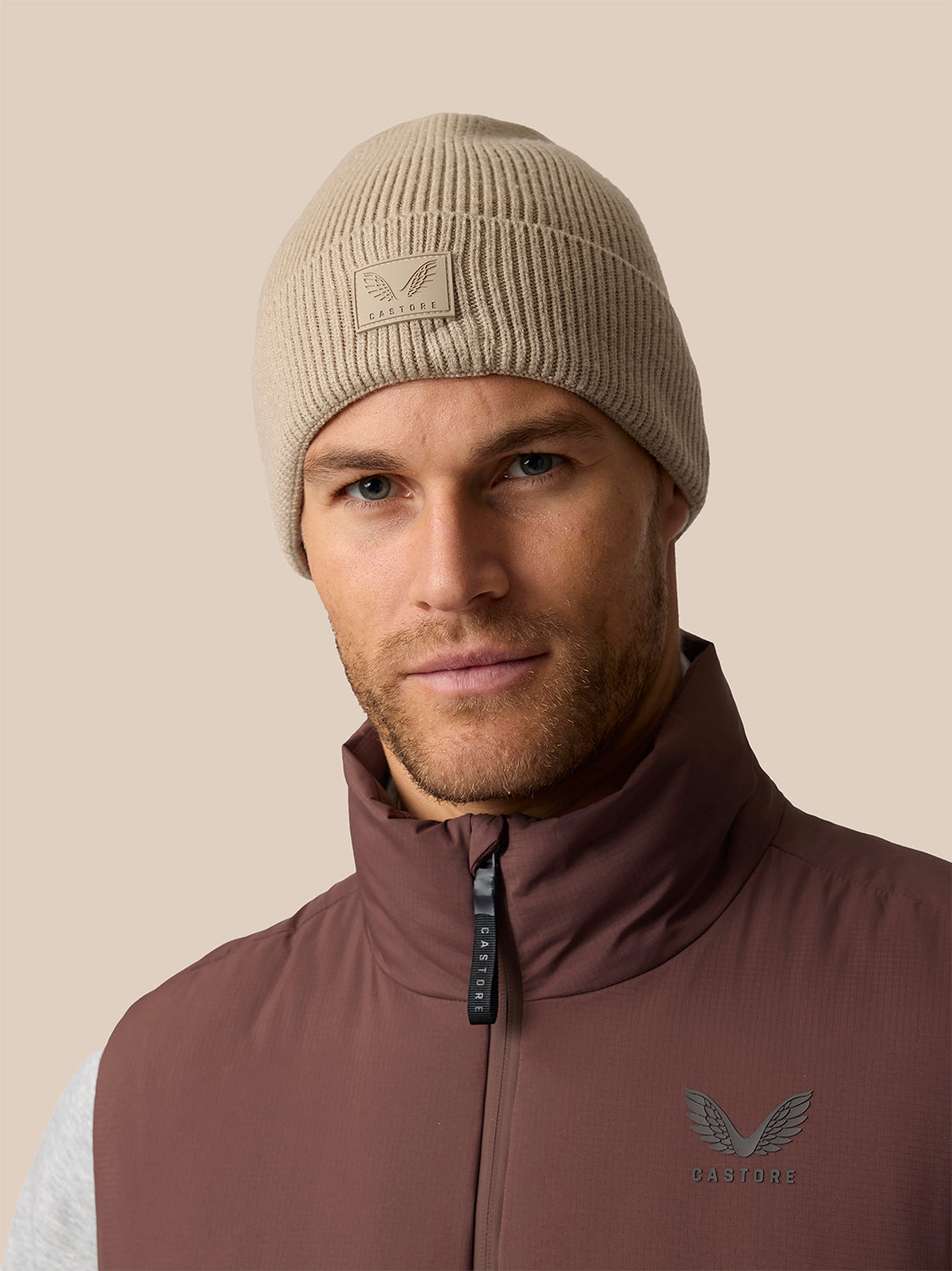 A man wearing a beige ribbed knit beanie with a winged logo patch and a brown zip-up vest with a matching winged logo on the chest, set against a solid beige background.