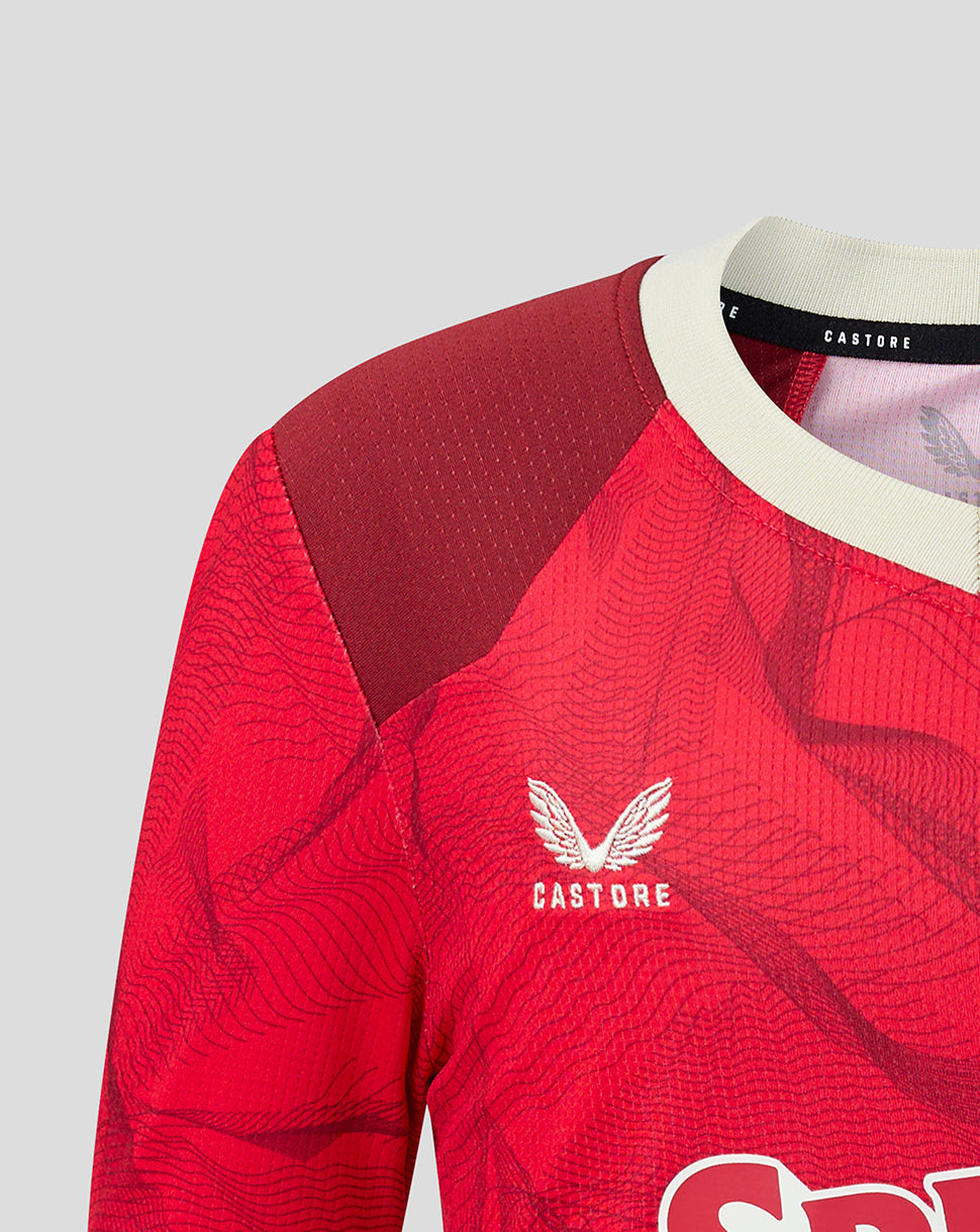 Close-up of a red Castore soccer jersey with a subtle wavy pattern, featuring a white wing logo and brand name on the chest, a cream collar, and a dark red textured shoulder panel.