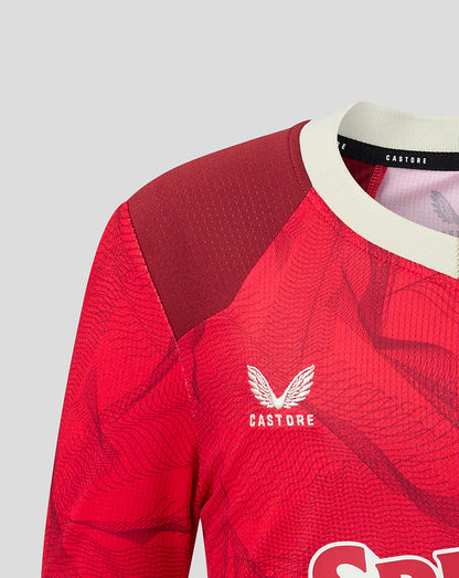 Close-up of a red Castore soccer jersey with a subtle wavy pattern, featuring a white wing logo and brand name on the chest, a cream collar, and a dark red textured shoulder panel.