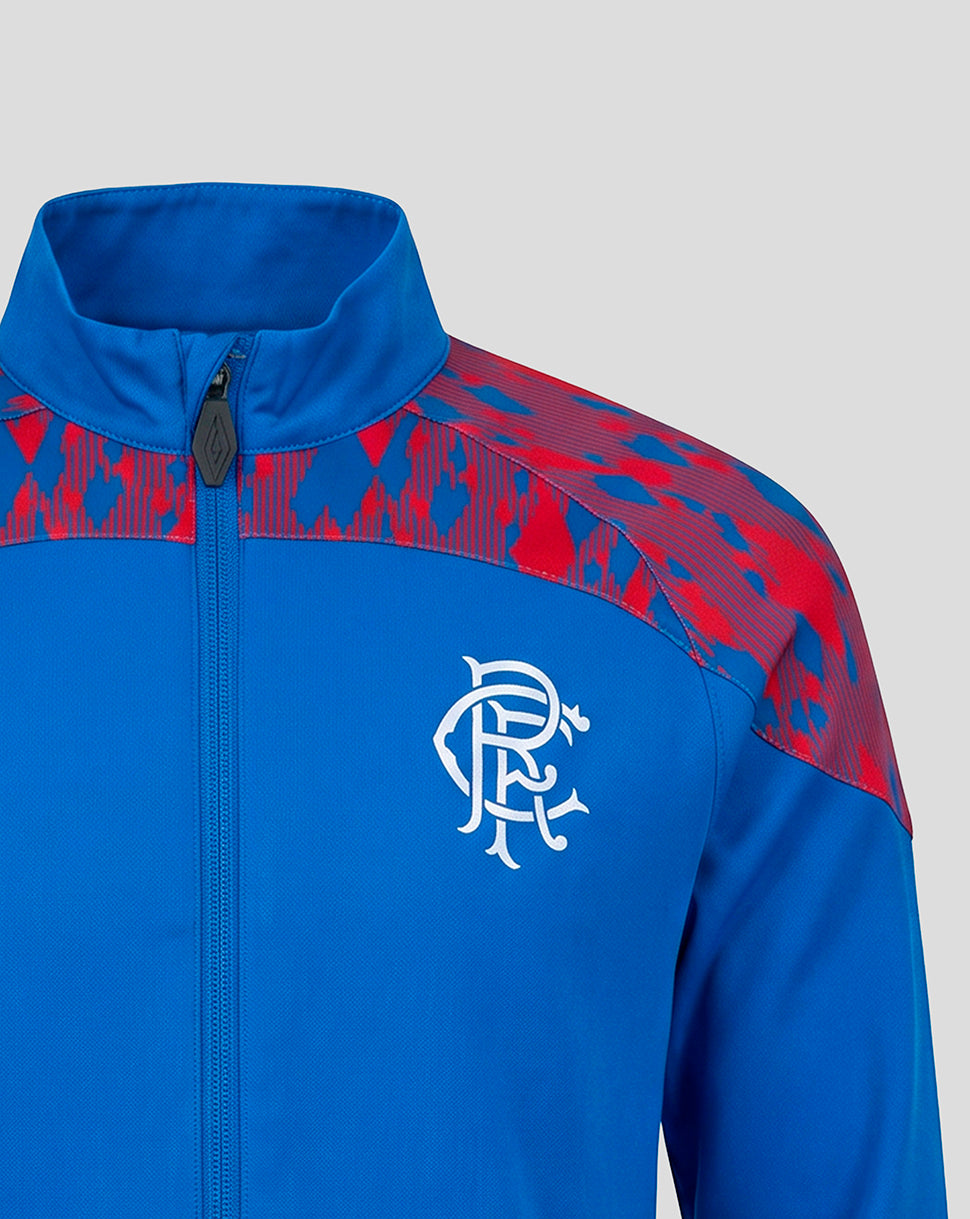 A close-up of a royal blue zip-up jacket with red patterned shoulder panels and a white Rangers FC crest on the chest, shown against a light gray background.