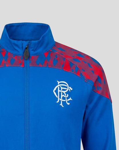 A close-up of a royal blue zip-up jacket with red patterned shoulder panels and a white Rangers FC crest on the chest, shown against a light gray background.
