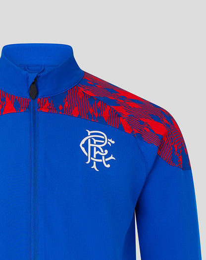A close-up of a royal blue athletic jacket featuring a red abstract patterned design across the shoulders and a white stylized crest logo on the chest, set against a solid light gray background.