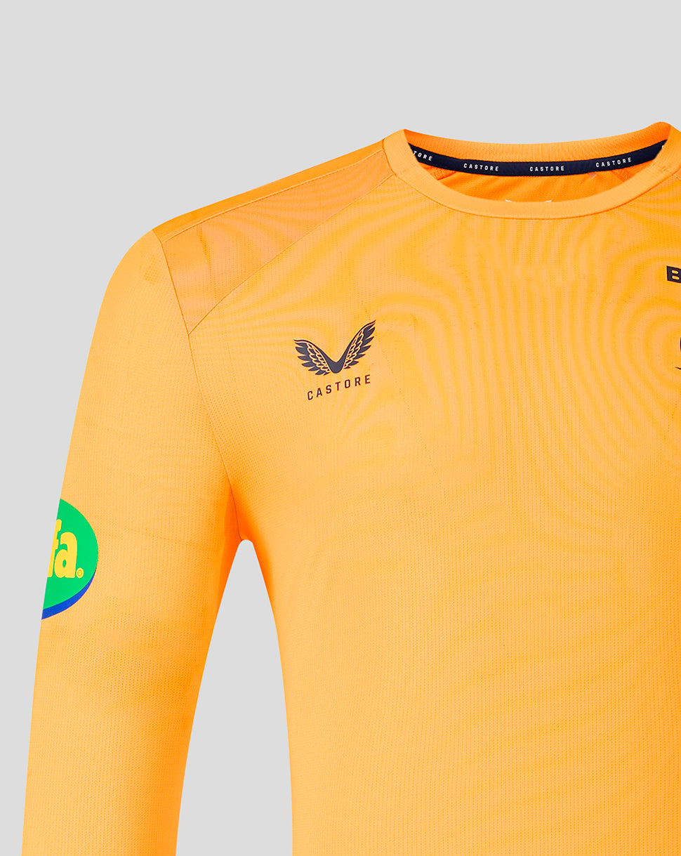 Close-up of a long-sleeve soccer jersey in bright orange with a subtle wavy texture, featuring the Castore wing logo on the chest and a green and blue sponsor patch on the sleeve, set against a light gray background.