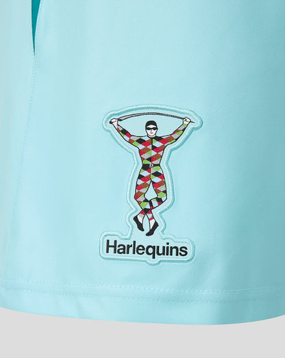A close-up of a light blue garment featuring a sewn-on patch with the Harlequins logo. The patch depicts a figure in a red, green, yellow, and black harlequin pattern holding a rope, with the word 'Harlequins' printed below in black text on a white background.