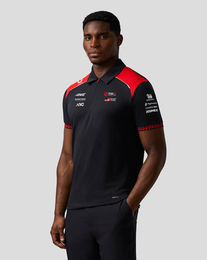 Man wearing a black and red polo shirt with various logos on a gray background