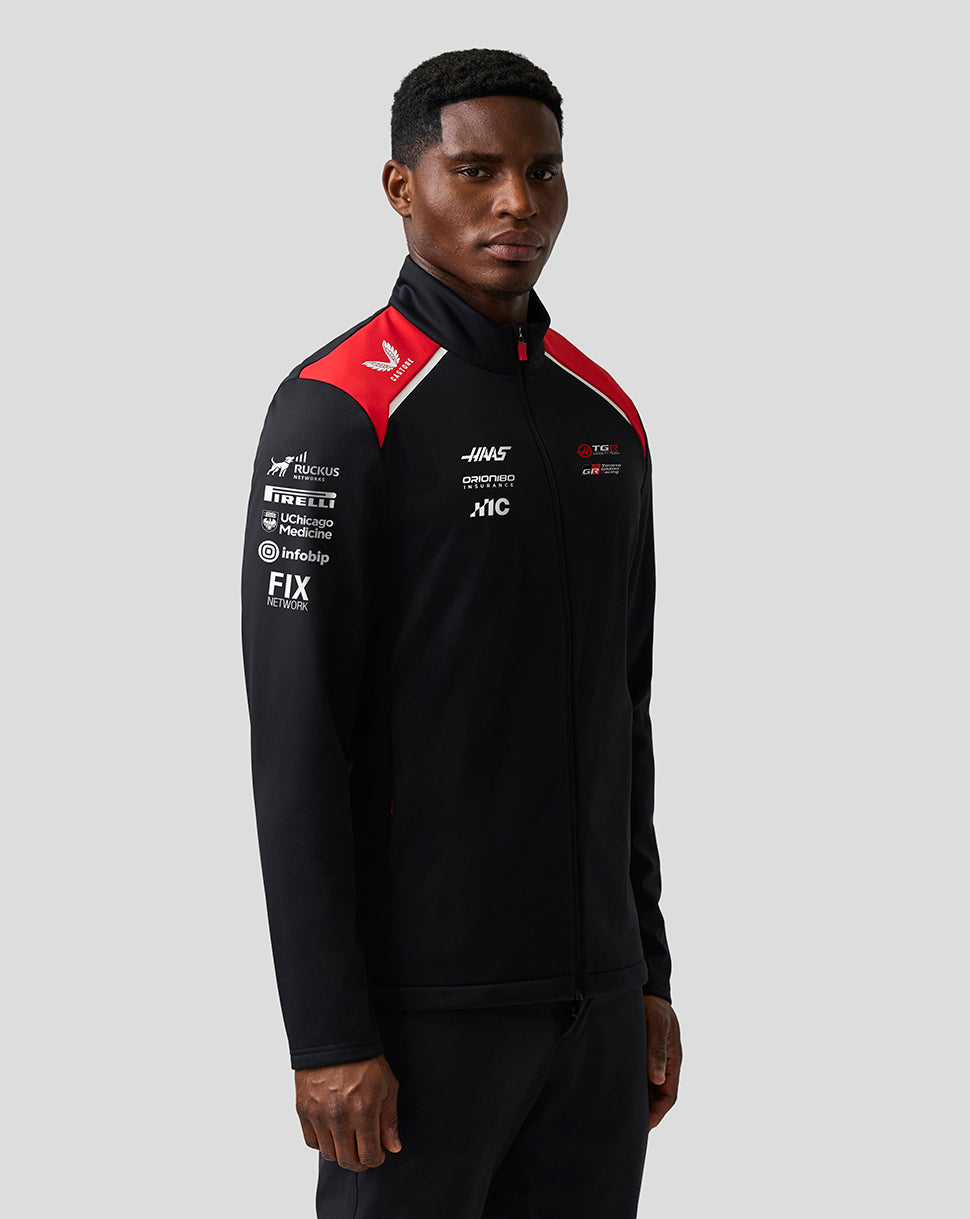 Person wearing a black and red sports jacket with various logos on a gray background