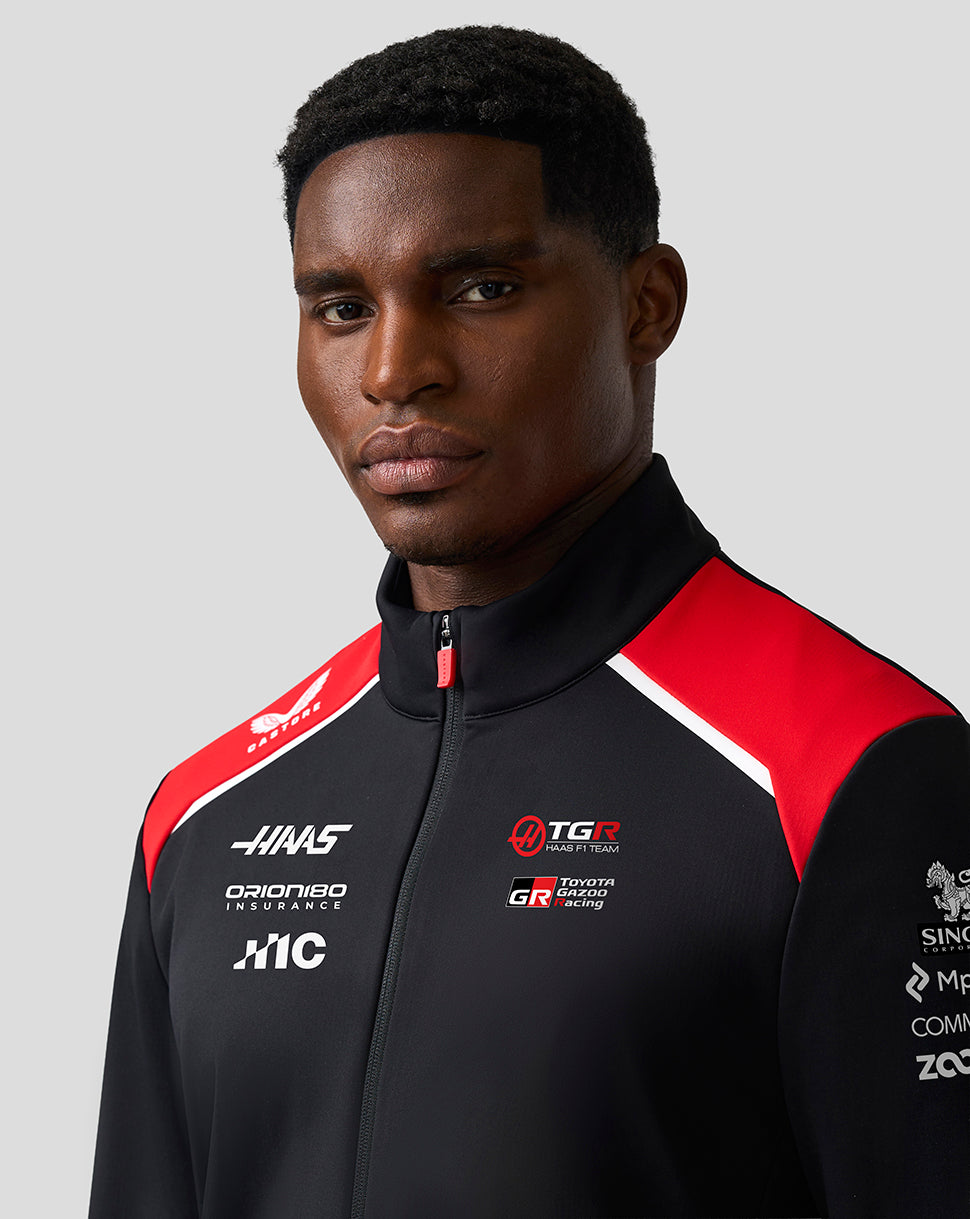 Man wearing a black and red jacket with various logos on a gray background
