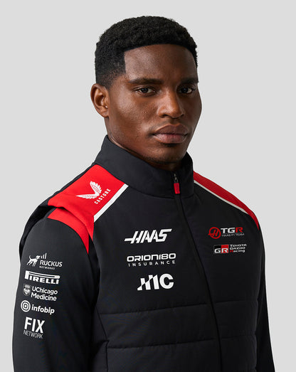 Person wearing a black racing jacket with various sponsor logos on a gray background