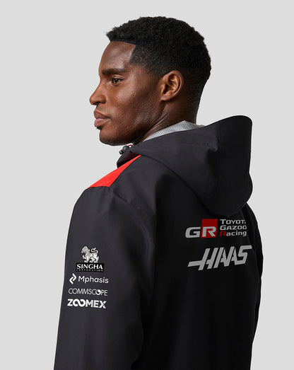 Person wearing a black jacket with various logos on the back against a gray background